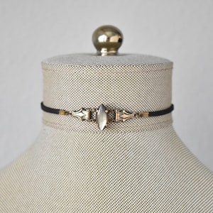 May include: A black choker necklace with an ornate, antique-style pendant. The pendant features a central, oval-shaped, clear stone and decorative metalwork in gold and silver tones. The necklace is displayed on a mannequin.