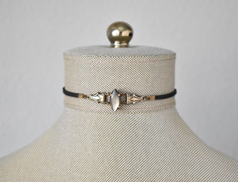 May include: A black choker necklace with an ornate, antique-style pendant. The pendant features a central, oval-shaped, clear stone and decorative metalwork in gold and silver tones. The necklace is displayed on a mannequin.
