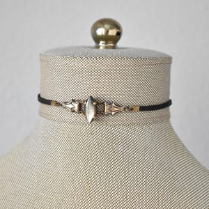 May include: A black choker necklace with a central silver-toned, elongated oval pendant. The pendant is flanked by ornate, gold-toned metalwork. The necklace is displayed on a mannequin head with a neutral fabric.