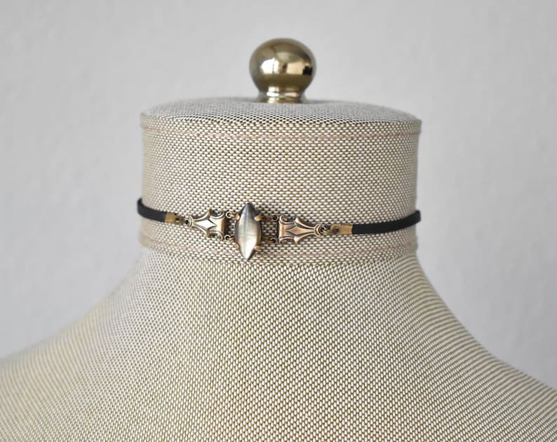 May include: A black choker necklace with a central silver-toned, elongated oval pendant. The pendant is flanked by ornate, gold-toned metalwork. The necklace is displayed on a mannequin head with a neutral fabric.