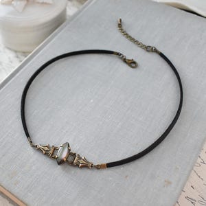May include: A black choker necklace with an ornate, antique-style centerpiece. The centerpiece features a pearlescent stone and bronze-colored metal accents. The necklace has a simple, elegant design, suitable for various occasions.