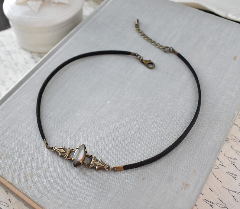 May include: A black choker necklace with an ornate, antique-style centerpiece. The centerpiece features a pearlescent stone and bronze-colored metal accents. The necklace has a simple, elegant design, suitable for various occasions.