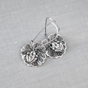 May include: Silver-toned frog earrings. Each earring features a detailed frog resting on a textured leaf. The earrings have a hook closure. The design is intricate and nature-inspired.