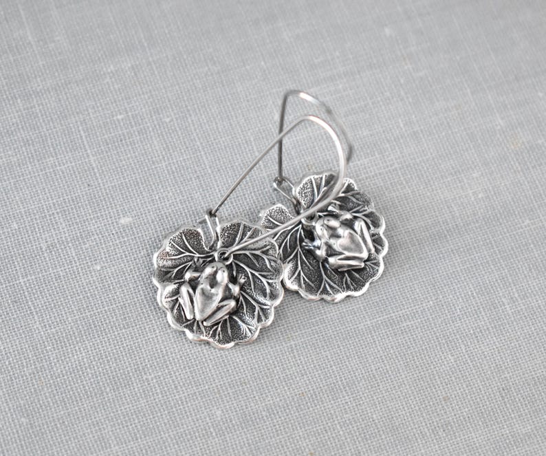 May include: Silver-toned frog earrings. Each earring features a detailed frog resting on a textured leaf. The earrings have a hook closure. The design is intricate and nature-inspired.
