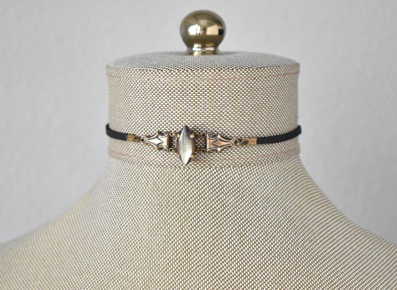 May include: A black choker necklace with an ornate silver-toned clasp and a central, elongated, oval-shaped stone. The necklace is displayed on a mannequin with a neutral-toned fabric.