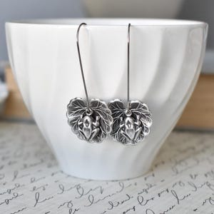May include: Silver-toned frog earrings. Each earring features a detailed frog charm resting on a leaf. The earrings hang from a simple wire hook. The background includes a white bowl and a patterned surface.