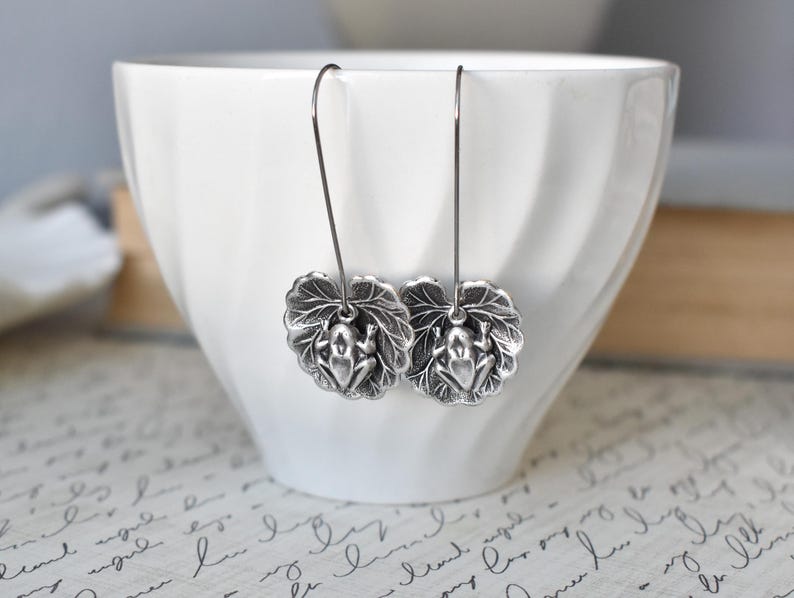 May include: Silver-toned frog earrings. Each earring features a detailed frog charm resting on a leaf. The earrings hang from a simple wire hook. The background includes a white bowl and a patterned surface.