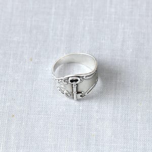 May include: A silver-toned ring with a key design. The ring features a detailed key motif at the center, with decorative flourishes around the band. The ring is set against a neutral background, highlighting its intricate design.