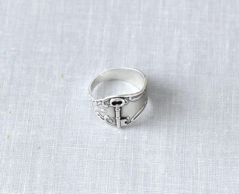 May include: A silver-toned ring with a key design. The ring features a detailed key motif at the center, with decorative flourishes around the band. The ring is set against a neutral background, highlighting its intricate design.
