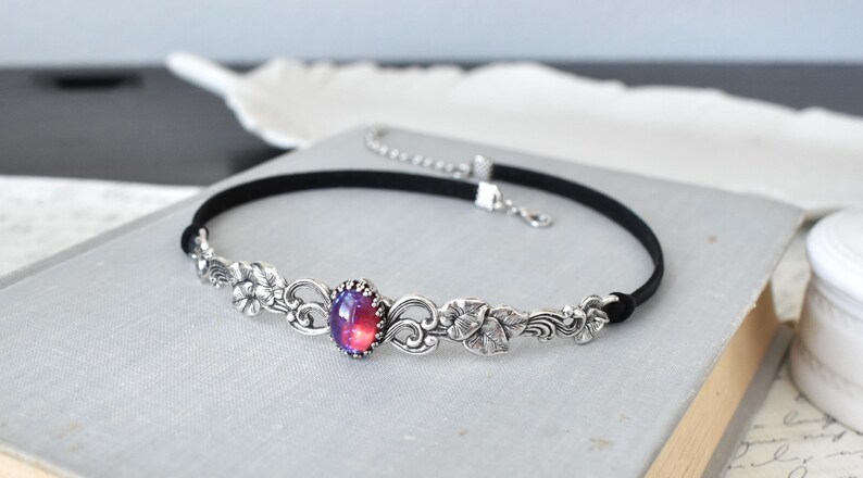 Puede incluir: Un collar gargantilla negro con un dise&ntilde;o floral plateado y una gran piedra preciosa roja y p&uacute;rpura iridiscente en el centro.