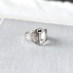 May include: A silver-toned ring with a key design. The ring is made from a spoon, with the key detail centered on the band. The ring has an antique, vintage aesthetic.