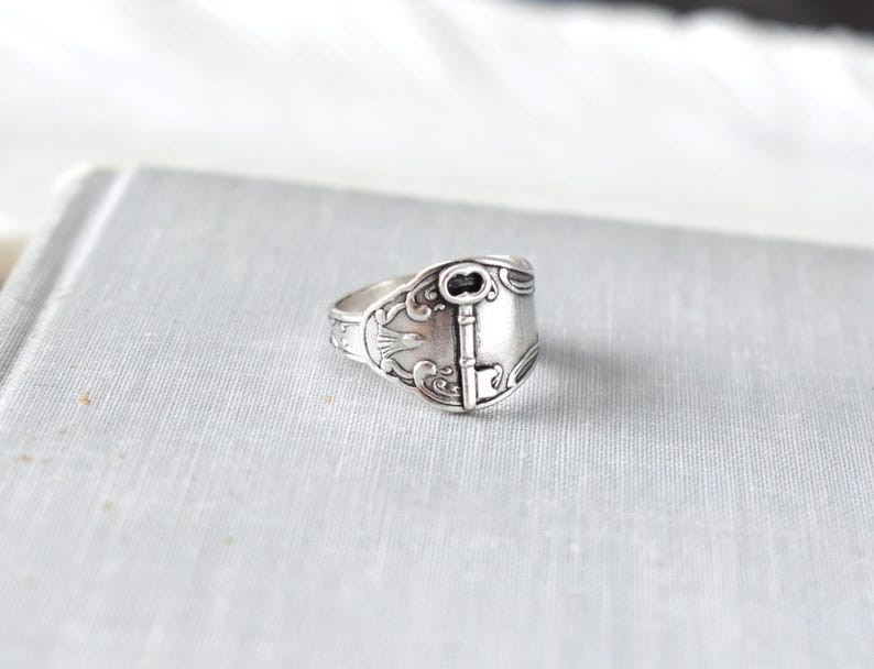 May include: A silver-toned ring with a key design. The ring is made from a spoon, with the key detail centered on the band. The ring has an antique, vintage aesthetic.
