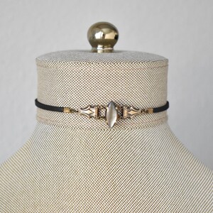 May include: A black choker necklace with a central, elongated, oval-shaped, clear stone. The necklace features ornate, antique-style metalwork in gold tones, and is displayed on a mannequin.