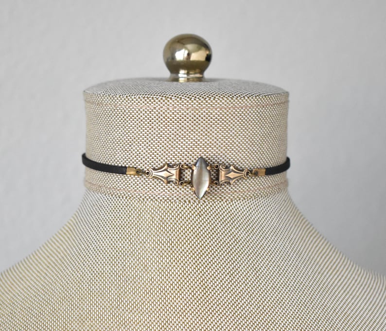 May include: A black choker necklace with a central, elongated, oval-shaped, clear stone. The necklace features ornate, antique-style metalwork in gold tones, and is displayed on a mannequin.