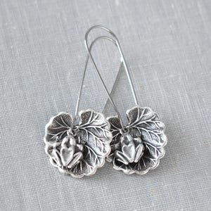 Peut inclure: Boucles d'oreilles argentées avec un motif de grenouille. Chaque boucle d'oreille présente un charm de grenouille détaillé posé sur un pendentif en forme de feuille. Les boucles d'oreilles sont suspendues à un simple crochet en fil de fer.