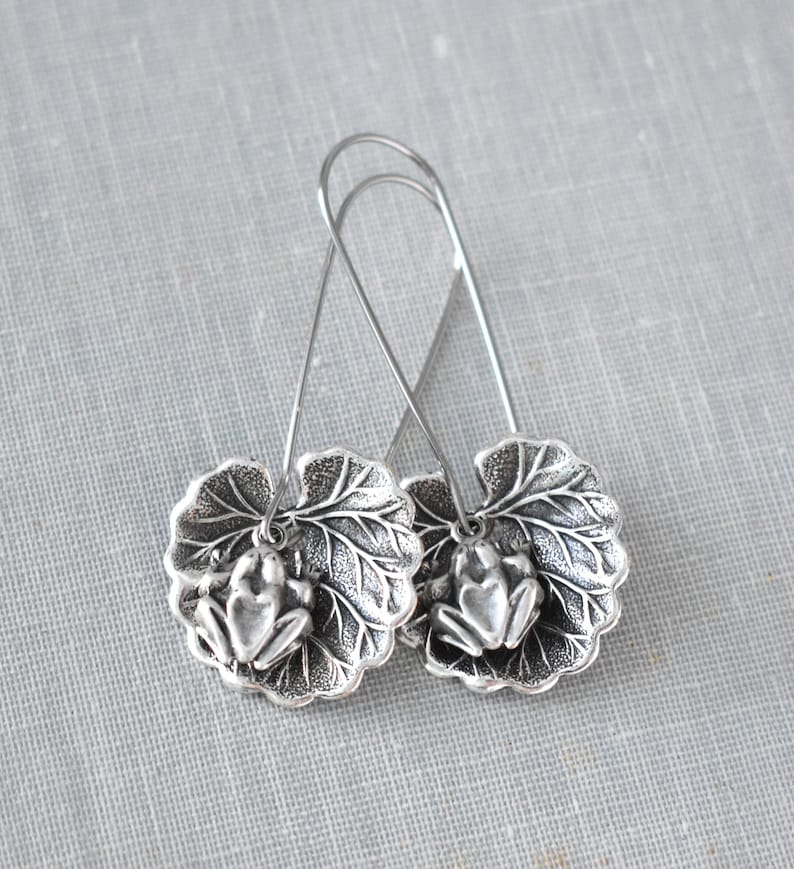 May include: Silver-toned earrings featuring a frog design. Each earring has a detailed frog charm resting on a leaf-shaped pendant. The earrings hang from a simple wire hook, creating a nature-inspired accessory.