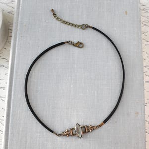 May include: A black choker necklace with an ornate, antique-style pendant. The pendant features a central, oval-shaped, iridescent stone set within a decorative, bronze-toned metal frame. The necklace has a clasp and an adjustable chain.
