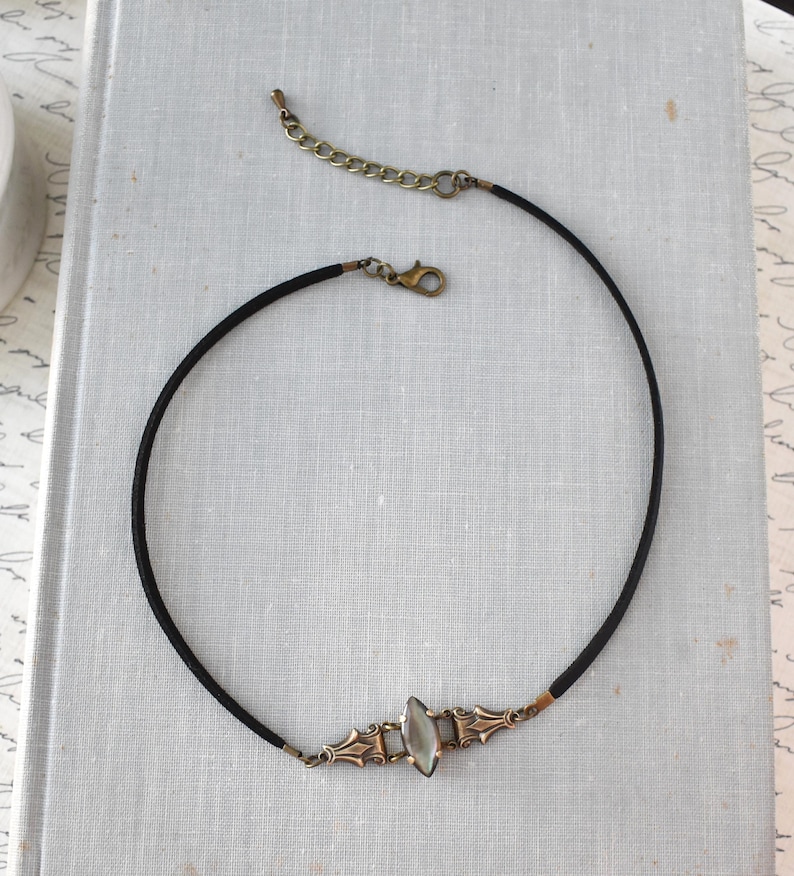 May include: A black choker necklace with an ornate, antique-style pendant. The pendant features a central, oval-shaped, iridescent stone set within a decorative, bronze-toned metal frame. The necklace has a clasp and an adjustable chain.