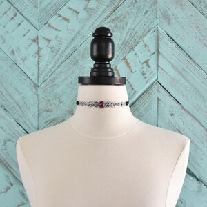 Puede incluir: Un collar choker plateado con un solo acento de piedra preciosa rosa. El collar se muestra en un maniqu&iacute; blanco sobre un fondo turquesa con patr&oacute;n de chevron.