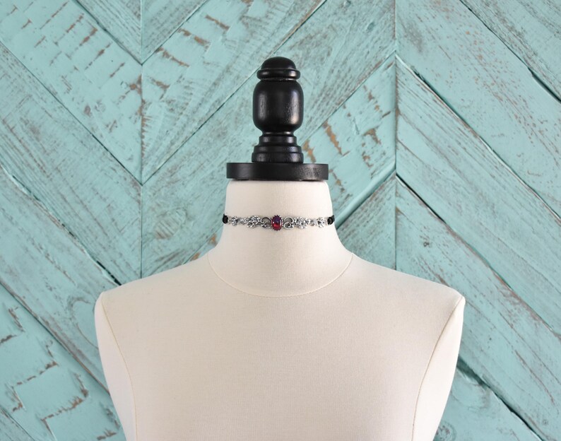 Puede incluir: Un collar choker plateado con un solo acento de piedra preciosa rosa. El collar se muestra en un maniqu&iacute; blanco sobre un fondo turquesa con patr&oacute;n de chevron.