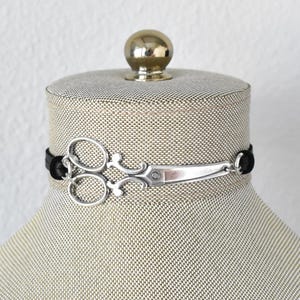 May include: A silver scissor charm choker necklace with a black cord. The scissors are detailed with a decorative design. The necklace is displayed on a mannequin head.