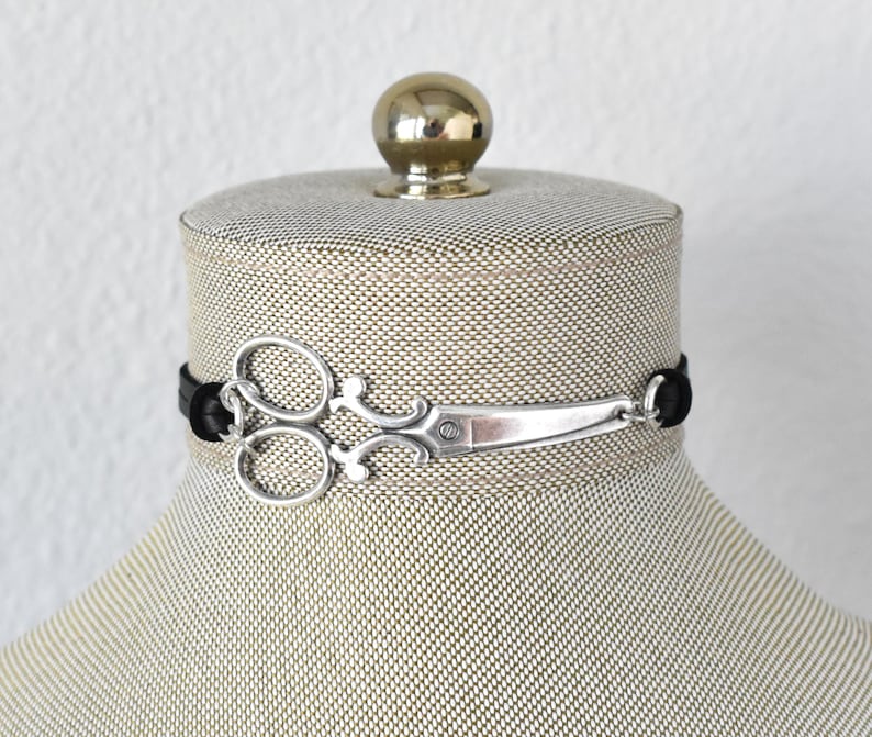 May include: A silver scissor charm choker necklace with a black cord. The scissors are detailed with a decorative design. The necklace is displayed on a mannequin head.