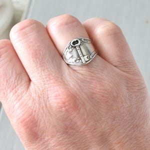 May include: A silver-toned spoon ring featuring a key design. The ring is worn on a hand, showcasing the intricate details of the key and floral embellishments. The ring's design is a unique piece of jewelry.
