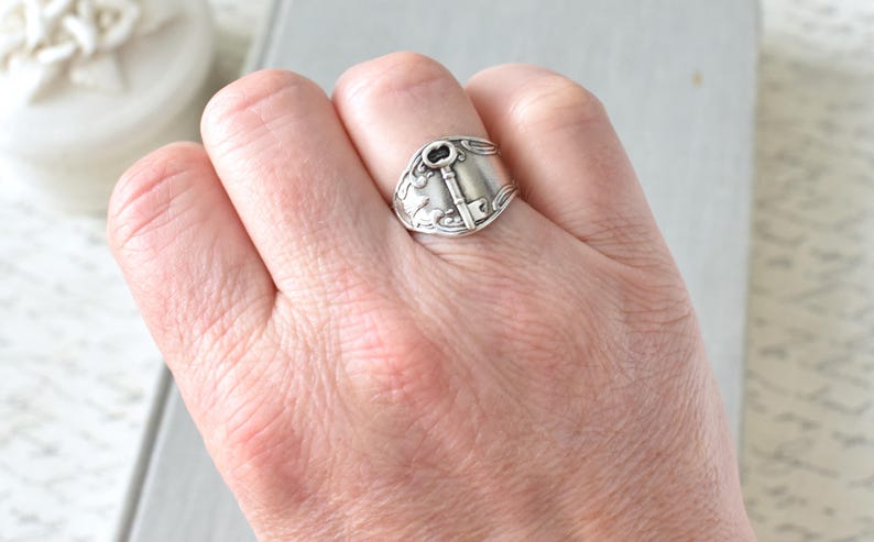 May include: A silver-toned spoon ring featuring a key design. The ring is worn on a hand, showcasing the intricate details of the key and floral embellishments. The ring's design is a unique piece of jewelry.