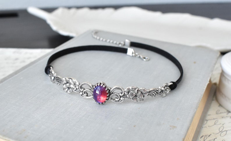 Puede incluir: Un collar choker plateado con un dise&ntilde;o floral y una gran piedra preciosa ovalada e iridiscente en el centro. El collar est&aacute; hecho de terciopelo negro y tiene un cierre plateado.