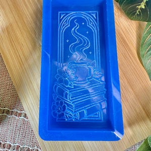 May include: A rectangular, bright blue silicone mold featuring an embossed design of a steaming mug atop a stack of books, framed by an arched window and flowers. The mold is designed for crafting decorative items.