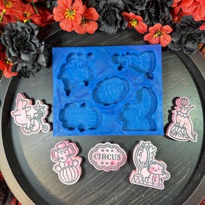 May include: A blue silicone mold featuring five distinct circus-themed cavities, accompanied by five corresponding pink and white resin pieces. The designs include a seal on a ball, a monkey on a bicycle, a tiger with a mirror, an elephant, and a sign reading CIRCUS.