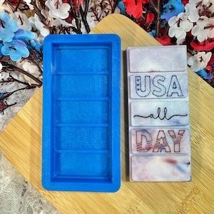 May include: A blue silicone mold and a rectangular, patriotic-themed resin piece. The resin piece has the words "USA all DAY" in red, white, and blue. The mold is empty and has the words "AQUA all DAY" imprinted.
