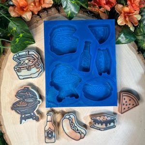 May include: A blue silicone mold featuring cavities shaped like a cooler, a grill, a bottle, a hot dog, a s'more, and a watermelon slice, accompanied by finished wax or soap pieces in the same shapes, including a grill, bottle, hot dog, s'more, and watermelon.