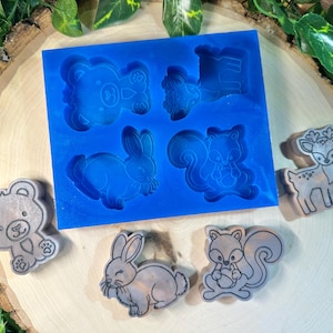 May include: A rectangular blue silicone mold featuring four distinct animal cavities: a bear, a deer, a rabbit, and a squirrel. Beside the mold are four corresponding finished figures, each showing detailed outlines of the bear, rabbit, squirrel, and deer.