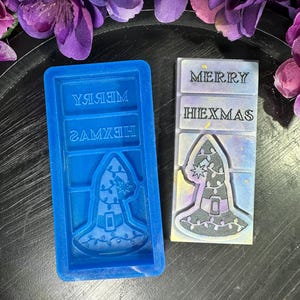 May include: A rectangular blue silicone mold and a matching wax melt bar. The mold features an embossed witch hat design and the text MERRY HEXMAS. The wax melt bar is white and purple with gold flecks, displaying the same witch hat and text.
