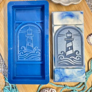 May include: A blue silicone mold and a finished soap bar featuring a lighthouse design. The soap bar has a blue and white marbled effect, with a lighthouse image in a rectangular frame. The mold is a vibrant blue, and the soap bar is on a wooden surface.