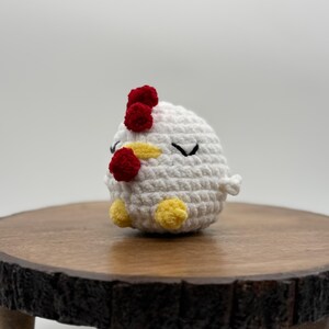 May include: A handmade, white crochet chicken with red comb and wattle, yellow feet, and black eyes. The small, round chicken is sitting on a wooden, round stand with a dark brown edge. The background is a plain white.