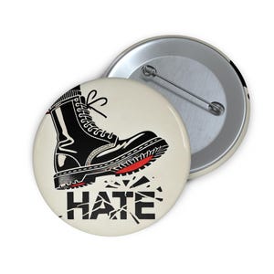 May include: A black and white button with a black boot stepping on the word "hate" which is broken into pieces. The boot has a red sole.