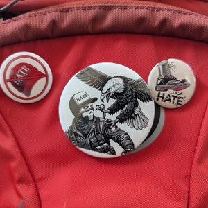 以下が含まれることがあります： 異なるデザインの丸いボタンピンが3つあります。最初のピンは、「Hate」と書かれた赤い野球帽です。2番目のピンは、黒いマスクと「Hate」と書かれた帽子をかぶった人物の白黒のイラストです。人物はハクトウワシを持っています。3番目のピンは、「Hate」と書かれた黒いブーツです。