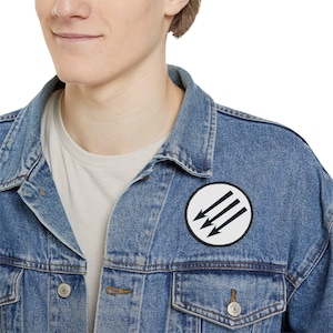 Puede incluir: Una chaqueta vaquera azul con un parche circular blanco que presenta tres flechas negras que apuntan hacia la izquierda.