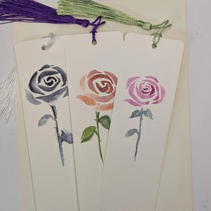 Può includere: Tre segnalibri a forma di rosa ad acquerello nei toni del grigio, marrone e rosa, ciascuno con uno stelo e foglie verdi. I segnalibri hanno nappe viola e verdi attaccate in alto. I segnalibri sono su uno sfondo color crema.