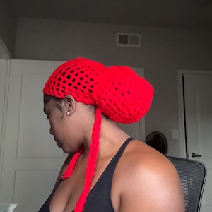May include: A red crocheted hat with a bun on top. The hat has long ties that hang down the front of the person wearing it.