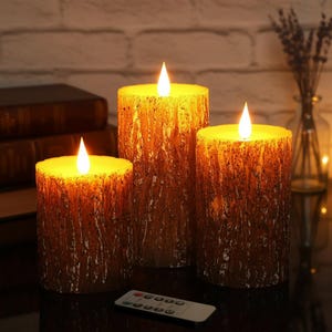 Peut inclure: Trois bougies LED cylindriques avec une finition extérieure texturée ressemblant à de l'écorce brune. Chaque bougie présente une flamme blanche chaude et lumineuse au sommet. Une petite télécommande rectangulaire est placée devant.