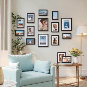 May include: A gallery wall arrangement featuring thirteen black picture frames of various sizes, each containing a photograph with a white mat board. The frames are displayed in a cluster on a light-coloured wall above a light blue armchair.