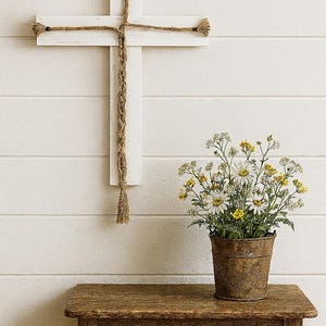 May include: A white wooden cross with rope detailing hangs on a white wall. A small, rustic wooden table holds a metal bucket filled with daisies and yellow flowers. The scene evokes a farmhouse aesthetic.
