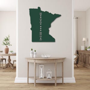 May include: A dark green metal wall art piece in the shape of the state of Minnesota. The art piece has a series of white dots and lines on the left side. The art is mounted on a white wall in a room with wooden floors and furniture.