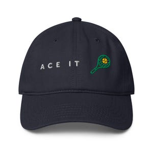 Peut inclure: Casquette de baseball bleu marine avec le texte "ACE IT" brodé en blanc. Un motif de raquette et de balle de tennis vert et jaune est brodé sur le côté droit. La casquette a une visière incurvée et une fermeture à sangle en tissu.