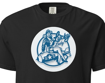 May include: Black t-shirt featuring a blue and white graphic of a woman sitting on a bomb, with the words "Lucky Lady" in a stylized font. The design is set within a white circle.