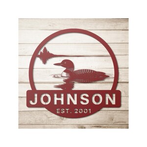 May include: A red metal wall art piece featuring a loon swimming in a lake, framed by a circle. The name "JOHNSON" and the year "EST. 2001" are displayed below the circle. The background is a light wood grain.