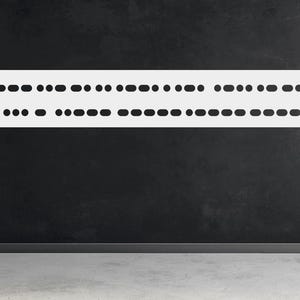 May include: A white, elongated wall decor piece with rounded ends, featuring a series of evenly spaced dots and dashes. The design is mounted on a dark gray wall above a light gray floor. The piece is a minimalist design.